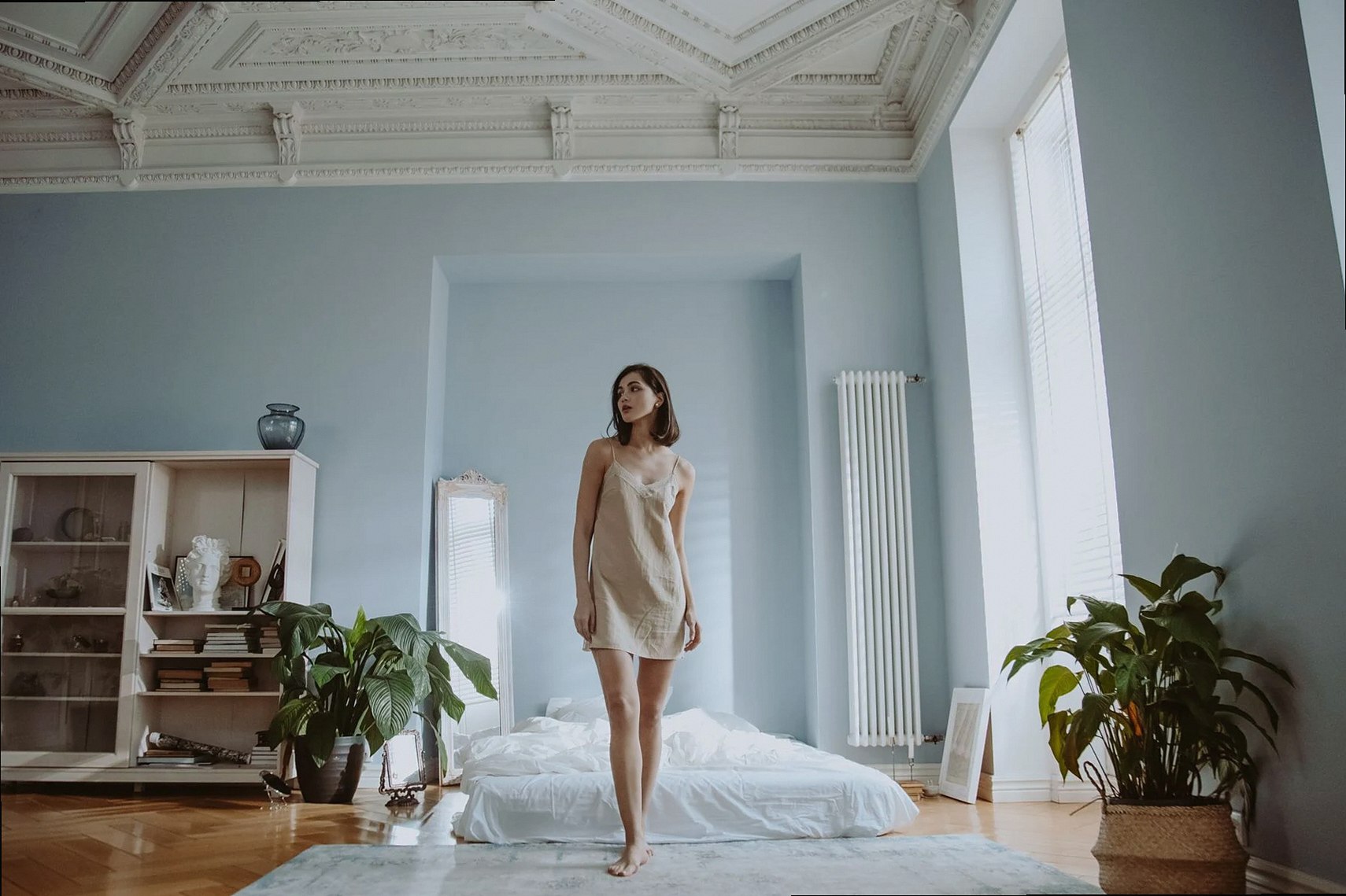 Elegant bedroom with a woman in nightwear, surrounded by houseplants and soft lighting. Cozy and serene setting.